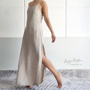 May include: A floor-length, light beige sleeveless dress with thin spaghetti straps, a scoop neckline, and a high side slit along the leg. The fabric has a visible natural texture. The text "bay bay" and "baybaylinen.com" appears in the corner.