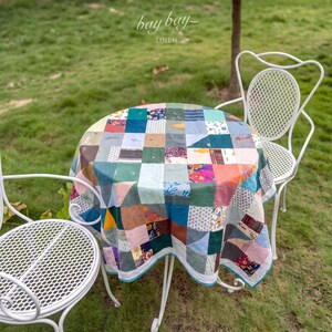 May include: A round table covered with a patchwork tablecloth featuring a variety of colorful fabric squares in shades of blue, green, pink, and yellow. Two white metal chairs with a lattice pattern are positioned on either side of the table.