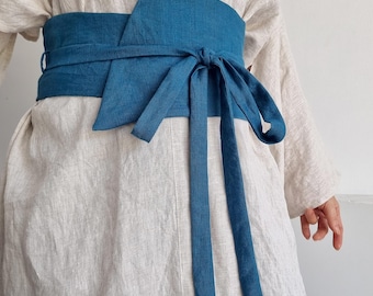 Peut inclure: Une large ceinture obi en lin bleu sarcelle nouée à la taille sur une tunique en lin beige à manches longues. La ceinture présente de longs liens en tissu et un design enveloppant structuré. Le texte bay bay baybaylinen.com est visible.