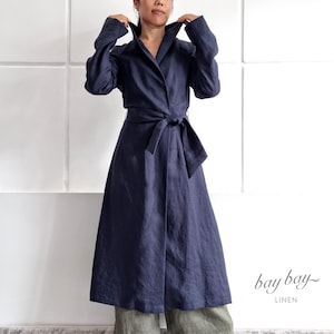 Peut inclure: Un manteau en lin bleu marine foncé à manches longues avec un large col, une ceinture à nouer à la taille et une jupe longue. Le manteau est associé à un pantalon large vert olive et des chaussures noires ouvertes. Le texte "bay bay LINEN" est visible.