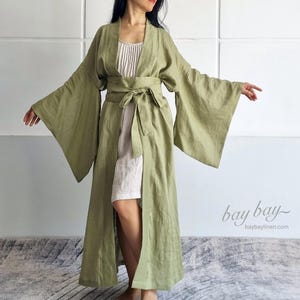 Puede incluir: Una bata larga de lino verde oliva con mangas anchas y cinturón a juego. La bata se lleva sobre un vestido de color claro. La imagen incluye el texto "bay bay" y "baybaylinen.com" en la esquina inferior derecha.