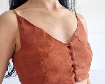 Peut inclure: Un haut court de couleur rouille avec un décolleté en V, de fines bretelles et une patte de boutonnage avant dotée de quatre boutons recouverts de tissu assorti. Le vêtement présente des coutures verticales et le texte bay bay LINEN.