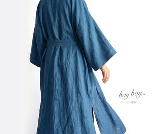 May include: A long-sleeved, calf-length blue linen robe featuring a tie-waist belt, wide sleeves, and side slits. The garment is shown from the back, and the text "bay bay LINEN" is printed in a dark font on the lower right side of the image.