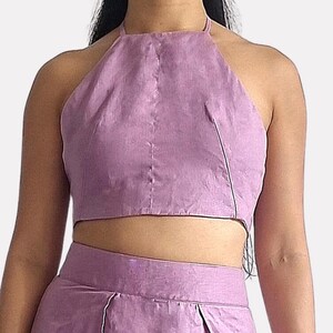 May include: A lavender linen two-piece set consisting of a cropped halter top with a high neckline and a matching skirt. The top features a central vertical seam and thin straps. The text "bay bay LINEN" is visible in the upper left corner.