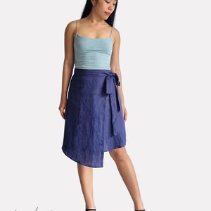 May include: A woman wearing a light blue tank top and a dark blue linen wrap skirt with a tie at the waist. She is wearing black heeled sandals. The skirt is asymmetrical and has a slightly uneven hemline.