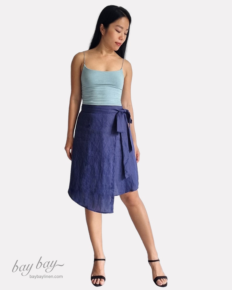 May include: A woman wearing a light blue tank top and a dark blue linen wrap skirt with a tie at the waist. She is wearing black heeled sandals. The skirt is asymmetrical and has a slightly uneven hemline.