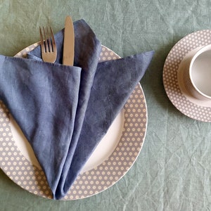 May include: A blue linen napkin folded into a triangle shape on a white plate with a gray and white polka dot pattern. A fork and knife are placed on top of the napkin.