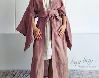 May include: A dusty rose, open-front long robe with wide, split sleeves and a matching tie-waist belt, worn over a cream-colored, sleeveless, pleated dress. The text 'bay bay' and 'baybaylinen.com' is visible in the lower right corner of the image.