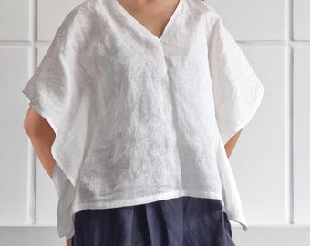 May include: A white linen top with a V-neckline and short, wide sleeves, paired with dark blue trousers. The top features a loose, boxy fit and a textured fabric finish. The text 'bay bay LINEN' is visible in the upper right corner of the image.