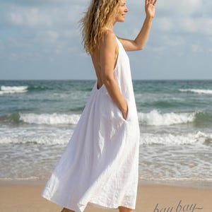 Puede incluir: Un vestido sin mangas de lino blanco con un diseño de espalda abierta, falda acampanada y bolsillos laterales. La prenda llega hasta los tobillos con una silueta sencilla, y el texto "bay bay LINEN" aparece en la esquina inferior derecha.