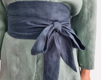 May include: A wide, dark blue linen sash belt tied in a bow at the front, worn over a light sage green linen garment. The belt features a textured fabric finish and long, tapered ends that drape downward from the central knot.