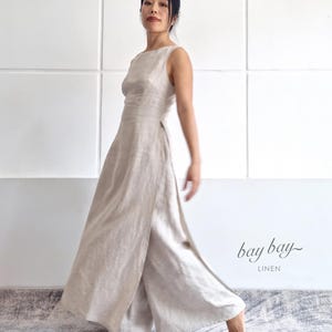 May include: A sleeveless beige linen jumpsuit featuring a high neckline and wide-leg trousers with a side slit. The garment is light-coloured and textured. The text "bay bay LINEN" is printed in the lower right corner of the image.