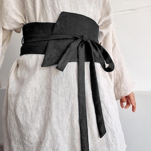 May include: A black obi belt tied around a cream-coloured linen garment. The wide belt wraps around the waist and ties in the front with long, flowing ties. The image includes the text "bay bay" and "baybaylinen.com" in the upper right corner.