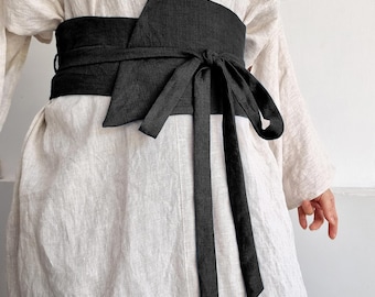 May include: A wide black wrap belt tied at the waist with long fabric straps, worn over a loose-fitting beige linen tunic. The text "bay bay" and "baybaylinen.com" is visible in the upper right corner of the image.