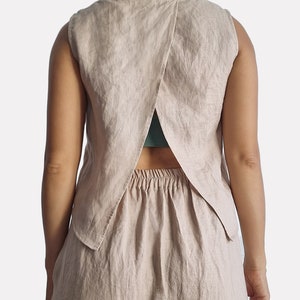 Può includere: Un top senza maniche in lino beige con un design a schiena incrociata. Il top ha una vestibilità rilassata e una lunghezza leggermente corta. La schiena del top è aperta, rivelando un reggiseno verde.