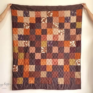 Puede incluir: Un edredón de retazos en tonos marrón, naranja y beige, con un borde marrón oscuro. El edredón está hecho de parches de tela cuadrados con varios diseños, incluidos diseños florales y geométricos. El edredón está sostenido por dos manos.