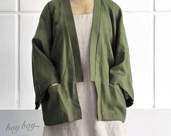 May include: An olive green open-front jacket with wide three-quarter length sleeves and two front patch pockets, worn over a light beige sleeveless dress. The text "bay bay LINEN" is visible in the lower left corner of the image.