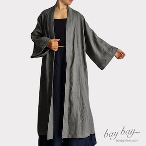 Puede incluir: Un abrigo largo tipo duster de color gris carbón, abierto por delante, con mangas anchas de tres cuartos y cuello estructurado. La prenda presenta un acabado de tela texturizada y llega hasta la pantorrilla. El texto bay bay baybaylinen.com es visible.