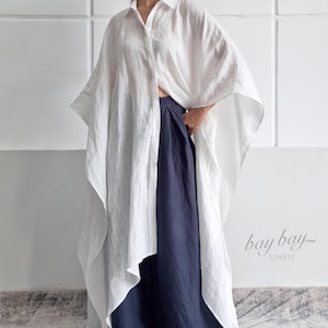 May include: A white oversized linen shirt with a classic collar and wide, flowing sleeves paired with dark navy blue wide-leg trousers. The garment features a button-down front and a loose, asymmetrical hemline. The text bay bay LINEN is visible.