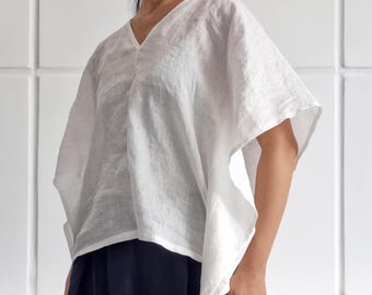 May include: A white linen top with a V-neck and short, wide sleeves. The top is paired with a dark blue skirt. The image shows a woman wearing the outfit. The brand name "bay bay~ LINEN" is visible in the bottom right corner.