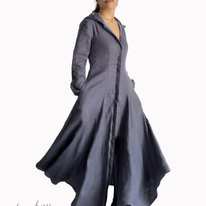 Puede incluir: Un abrigo largo de color gris pizarra con mangas largas, capucha, cierre de botones frontal y una falda acampanada y fluida. La prenda tiene un corpiño ajustado. El texto "bay bay" y "baybaylinen.com" está impreso en la esquina inferior izquierda.