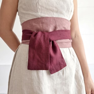 Puede incluir: Un vestido de lino sin mangas con una faja en capas burdeos y rosa atada con un lazo. El vestido es de color beige claro, y la faja añade un toque de color. El texto "bay bay~ LINEN" es visible en la parte inferior derecha.