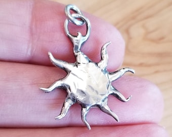 May include: Silver charm pendant in the shape of a sun with a textured surface and a loop for attaching to a chain or bracelet.