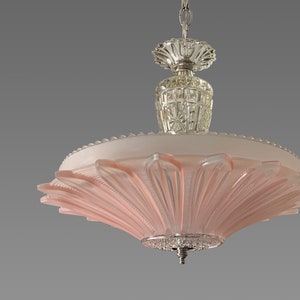 May include: A ceiling light fixture featuring a wide, pale pink glass shade with a radial, petal-like pattern. The fixture includes a clear, faceted glass central column, a chrome-finished canopy, and a small, decorative silver-toned finial at the base.