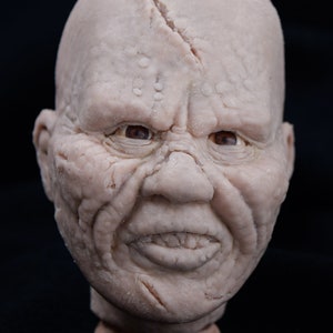 May include: A detailed, realistic sculpture of a human head with a wrinkled, textured face. The head has a large, prominent nose and a wide mouth with visible teeth. The sculpture is made of a light-colored material, possibly clay or plaster.
