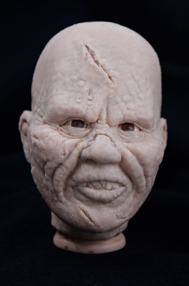 May include: A detailed, realistic sculpture of a human head with a wrinkled, textured face. The head has a large, prominent nose and a wide mouth with visible teeth. The sculpture is made of a light-colored material, possibly clay or plaster.