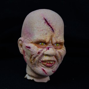 May include: A close-up of a grotesque, pale-skinned, sculpted head with yellow eyes, a wide, toothy grin, and red, bloody cuts and scrapes across the face.