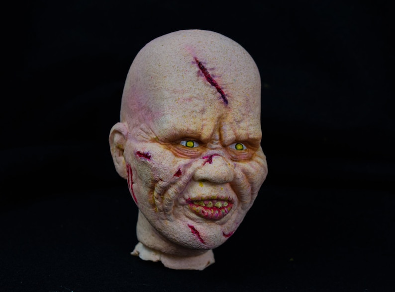 May include: A close-up of a grotesque, pale-skinned, sculpted head with yellow eyes, a wide, toothy grin, and red, bloody cuts and scrapes across the face.