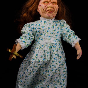 May include: A porcelain doll with red hair, a white dress with blue floral print, and a distressed face. The doll is holding a small wooden cross with a gold figure.