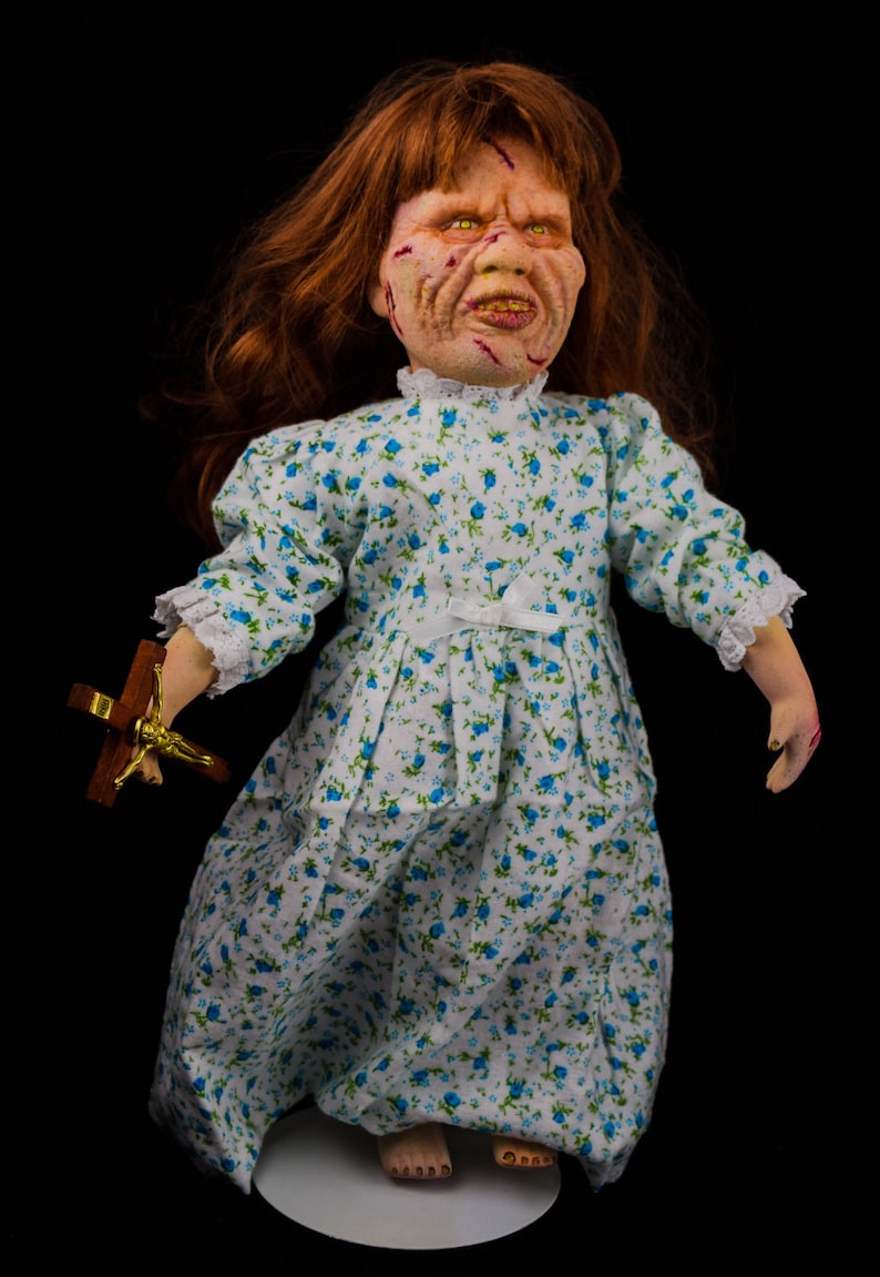 May include: A porcelain doll with red hair, a white dress with blue floral print, and a distressed face. The doll is holding a small wooden cross with a gold figure.