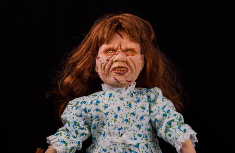May include: A creepy porcelain doll with red hair, wearing a white dress with blue floral print. The doll has a realistic face with scars and a sinister expression.