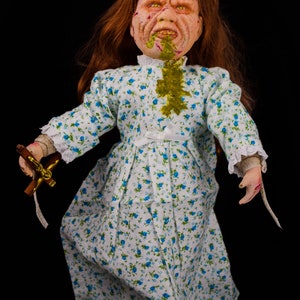 May include: A porcelain doll with long red hair, wearing a white dress with blue floral print. The doll has a grotesque expression with green slime on its face and a cross in its hand.