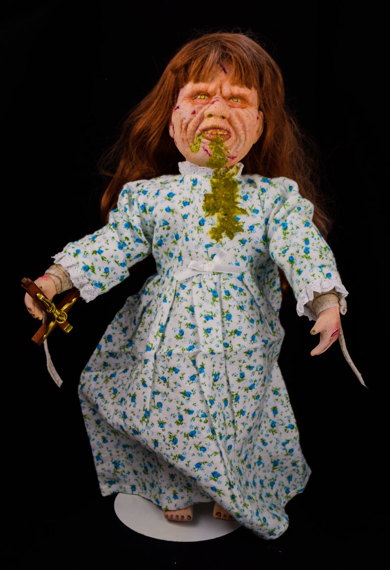 May include: A porcelain doll with long red hair, wearing a white dress with blue floral print. The doll has a grotesque expression with green slime on its face and a cross in its hand.