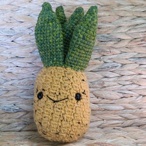 May include: A handmade crochet pineapple toy with a smiling face. The pineapple has a tan body with black speckles and green leaves. The toy is approximately 5 inches tall.