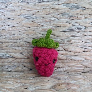 May include: A handmade, crocheted strawberry with a smiling face. The strawberry is a vibrant pink with a green leafy top and black eyes. The amigurumi strawberry is a cute, whimsical design.