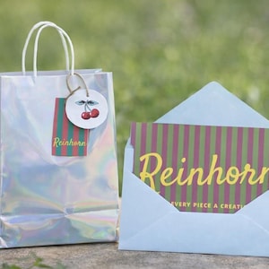 Pode incluir: Uma sacola de presente holográfica com alças brancas e uma etiqueta com cerejas e o nome "Reinhorn". Um cartão com o nome "Reinhorn" em escrita amarela está em um envelope azul claro. O cartão tem um padrão listrado.