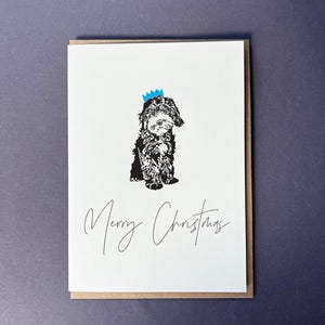 Può includere: Un biglietto di Natale con un'illustrazione in bianco e nero di un cane con una corona blu. Le parole "Merry Christmas" sono scritte in corsivo sotto l'illustrazione del cane. Il biglietto ha un fronte bianco e un bordo marrone.