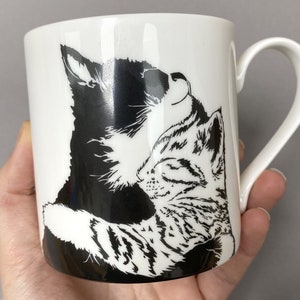 May include: White ceramic mug with a black and white silhouette of two cats cuddling. The cats are facing each other with their heads tilted back.