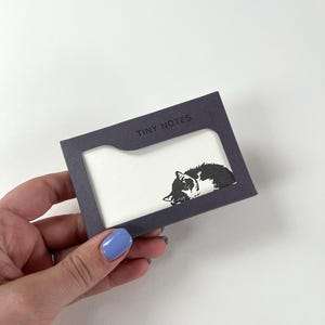 Könnte beinhalten: Eine dunkelgraue Karte mit dem Aufdruck "TINY NOTES" oben. Die Karte hat einen rechteckigen Ausschnitt mit einer schwarz-weißen Illustration einer schlafenden Katze. Die Karte wird von einer Person mit blauem Nagellack gehalten.