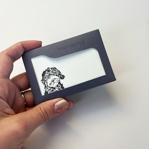 Pode incluir: Um bloco de notas azul escuro com as palavras "TINY NOTES" impressas no topo. O bloco de notas apresenta uma ilustração a preto e branco de um cão. O bloco de notas é segurado por uma mão.
