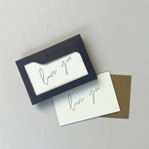 Peut inclure: Un ensemble de petits cartons avec un support bleu foncé. Le support a une fenêtre rectangulaire révélant une carte blanche avec la phrase manuscrite "love you". Une carte blanche séparée et une carte marron sont également visibles.