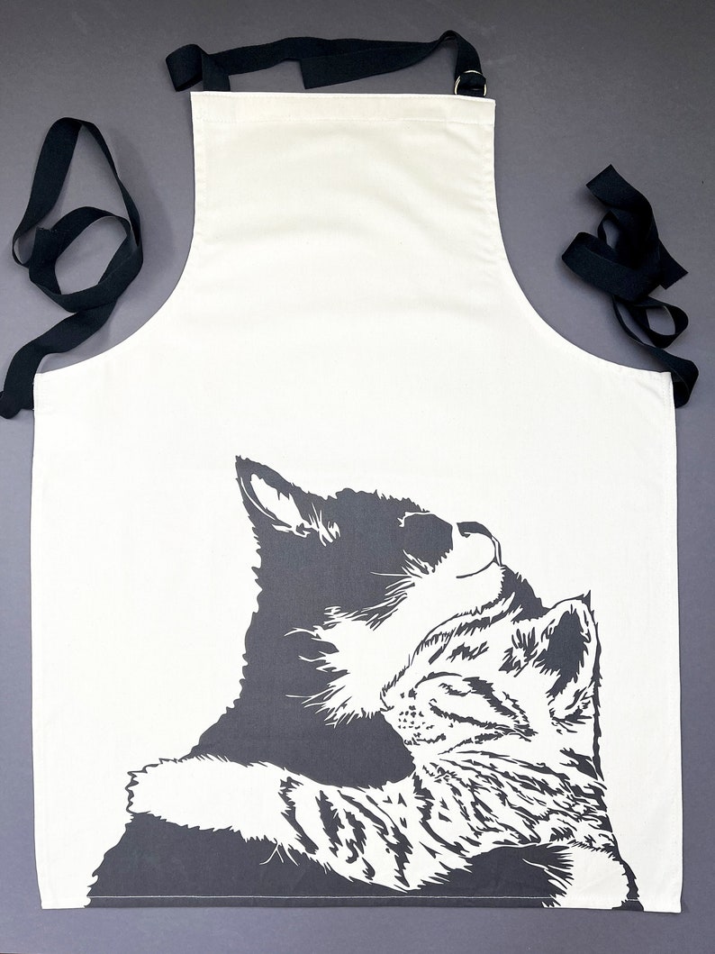 Lovely unbleached cotton apron with the cuddling cats illustration on the front with the image off the bottom. The apron is cream with black tie's and a black adjustable neck and brass fastening.