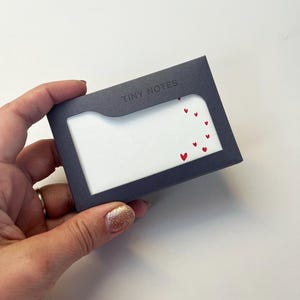 Op de afbeelding: Een kleine, donkergrijze kaarthouder met het opschrift "TINY NOTES" en een rechthoekig venster. Binnenin het venster is wit papier versierd met kleine rode hartjes. De kaarthouder wordt in een hand gehouden.