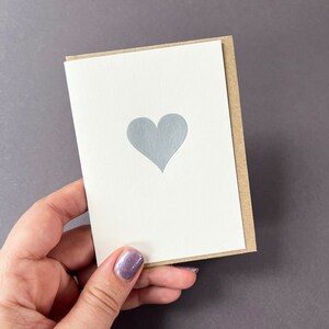 May include: A greeting card featuring a silver heart design on a white card, with a kraft paper backing. The card is held in a hand, against a grey background.