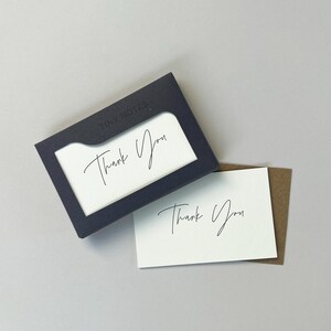 May include: A set of "Thank You" cards with a navy blue holder. The cards are white with elegant script lettering. The holder has the words "Tiny Notes" printed on it.