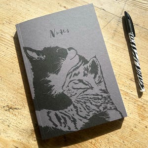 Könnte beinhalten: Ein graues Notizbuch mit dem Wort "Notes" und einer schwarzen Illustration von zwei Katzen. Ein schwarz-weiß gestreifter Stift liegt auf einer Holzoberfläche neben dem Notizbuch.